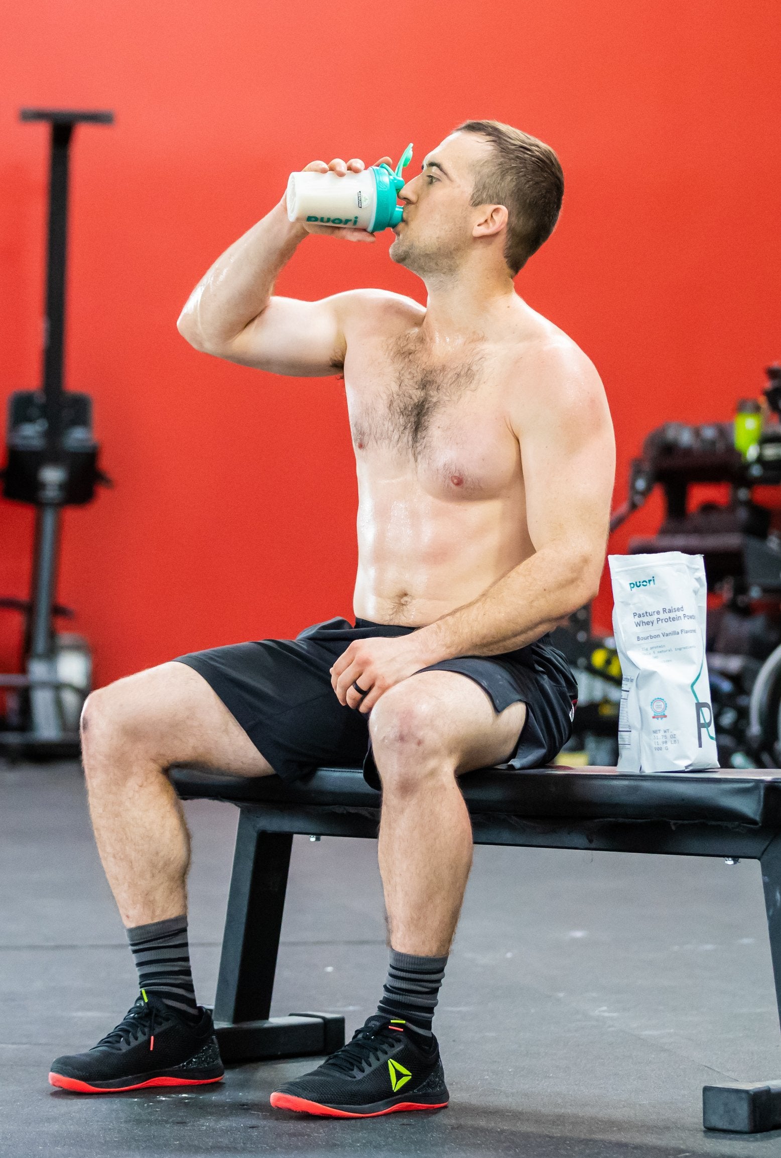 Shirtless man sitting on a gym bench drinking from a teal Puori shaker bottle, with a Puori whey protein bag on the bench beside him.